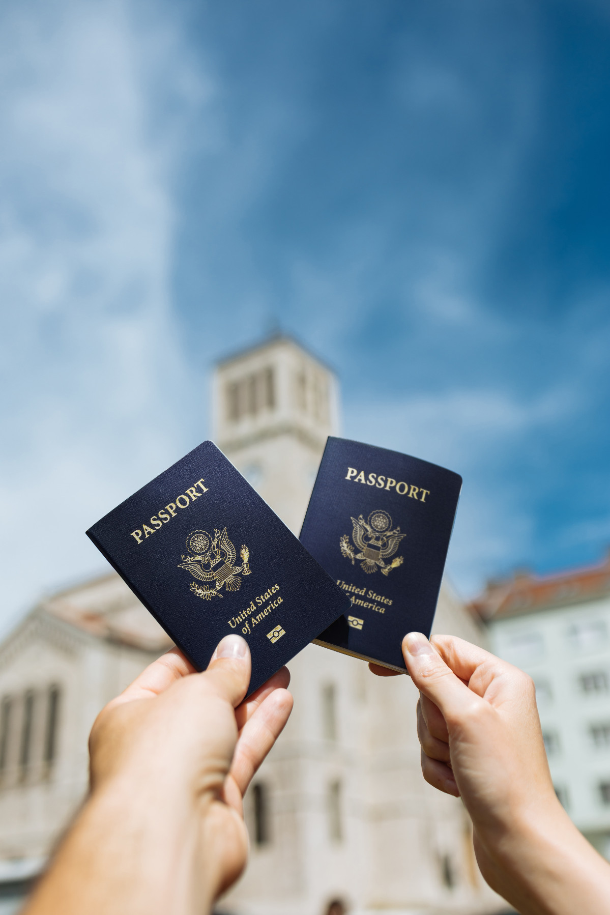 Two people's hands holding US passports against backdrop of historical buildings and blue sky